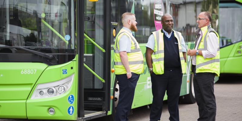 Three men in yellow high viz jackets stand talking in front of a green bus