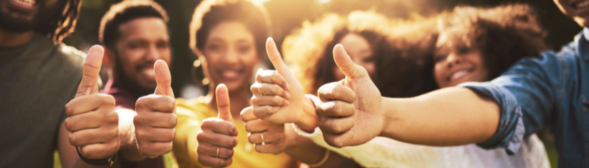 A group of people stand outdoors in a sunlit, natural setting, all giving a thumbs-up gesture toward the camera.