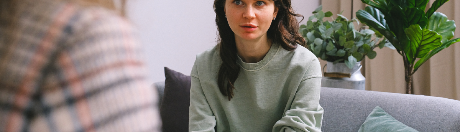 Women sat on a sofa having a conversation with someone. This other person's is facing away from the camera and only their shoulder and back of their head can be seen