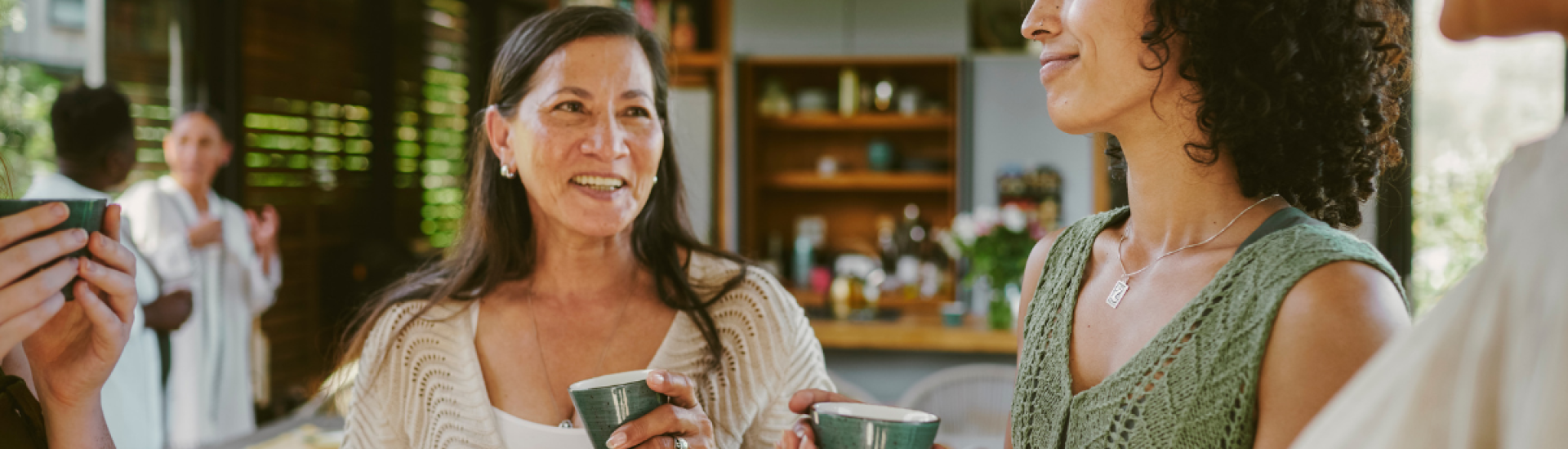 Several people gathered indoors in a modern kitchen setting, holding green ceramic cups. The background shows wooden shelves with various kitchen items, including bowls, jars, and plants.
