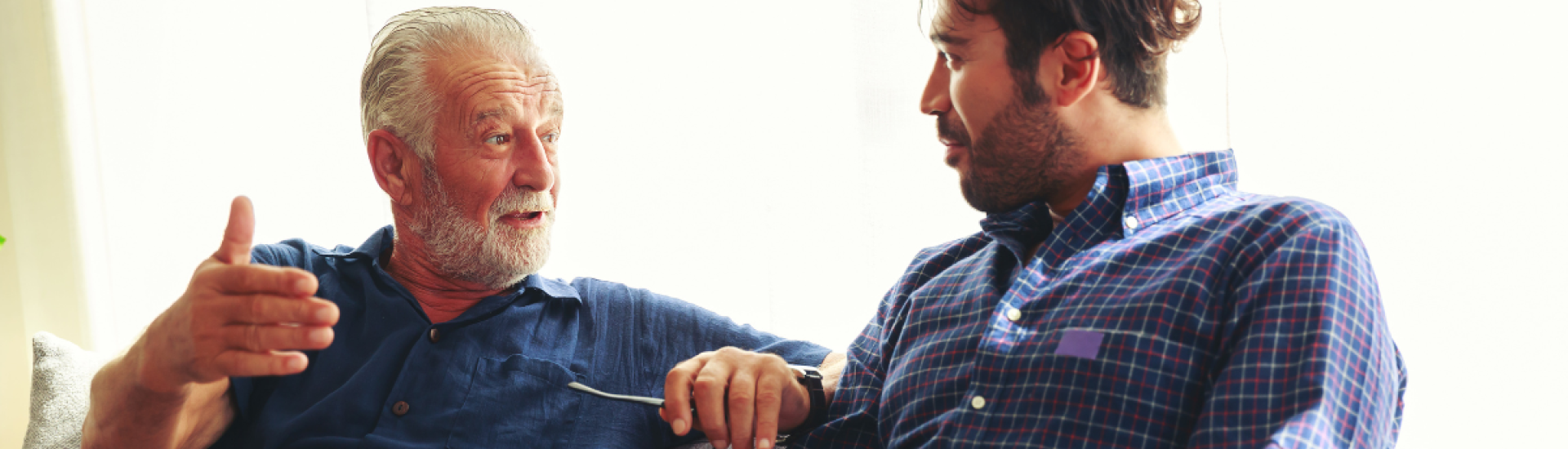 Two people sitting together on a sofa having a conversation. One gestures with his hand while holding a pair of glasses, and the other listens attentively. Soft natural light comes through a window behind them, creating a calm, informal setting.