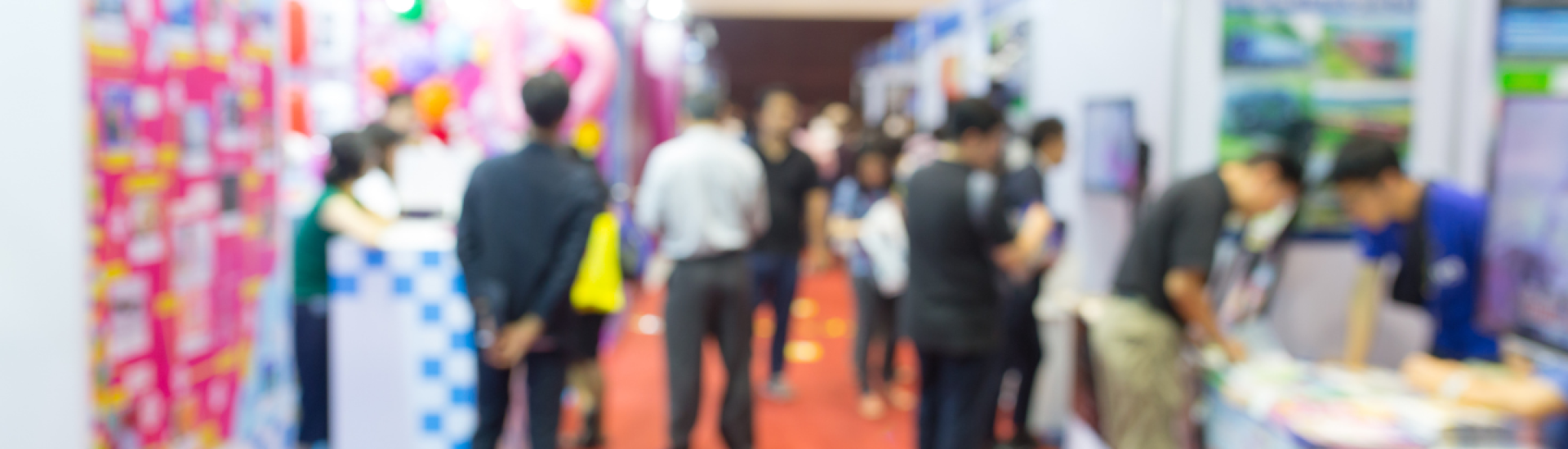 Blurred photo of an event with exhibiting stalls and individuals in conversation and walking around the event space