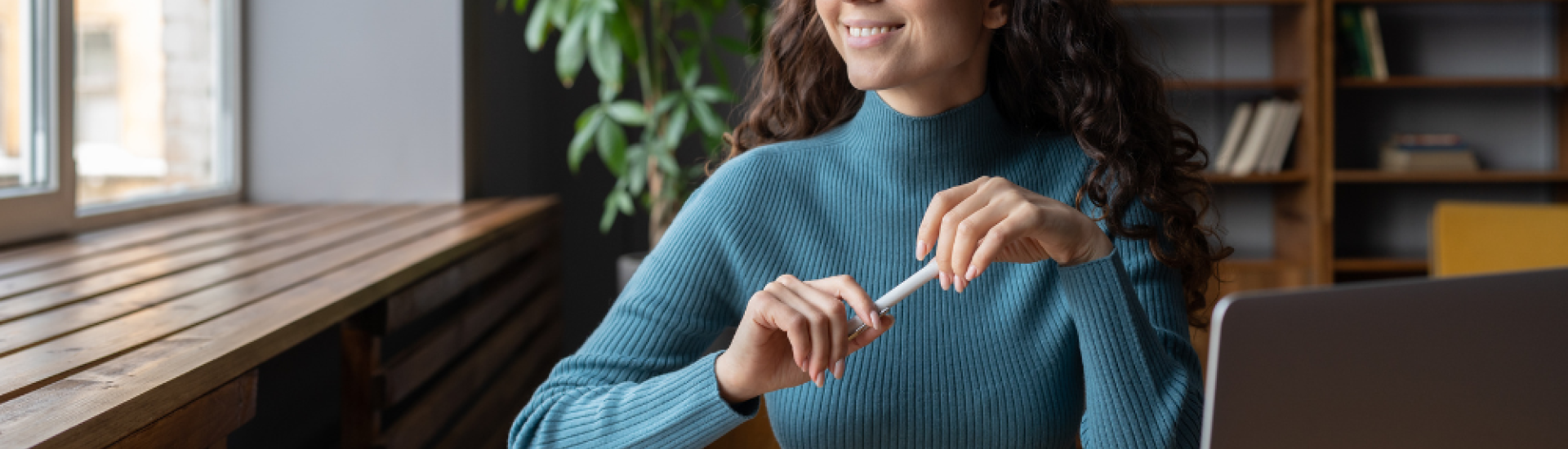 A woman looking out the window whilst sat at a desk and holding a pen