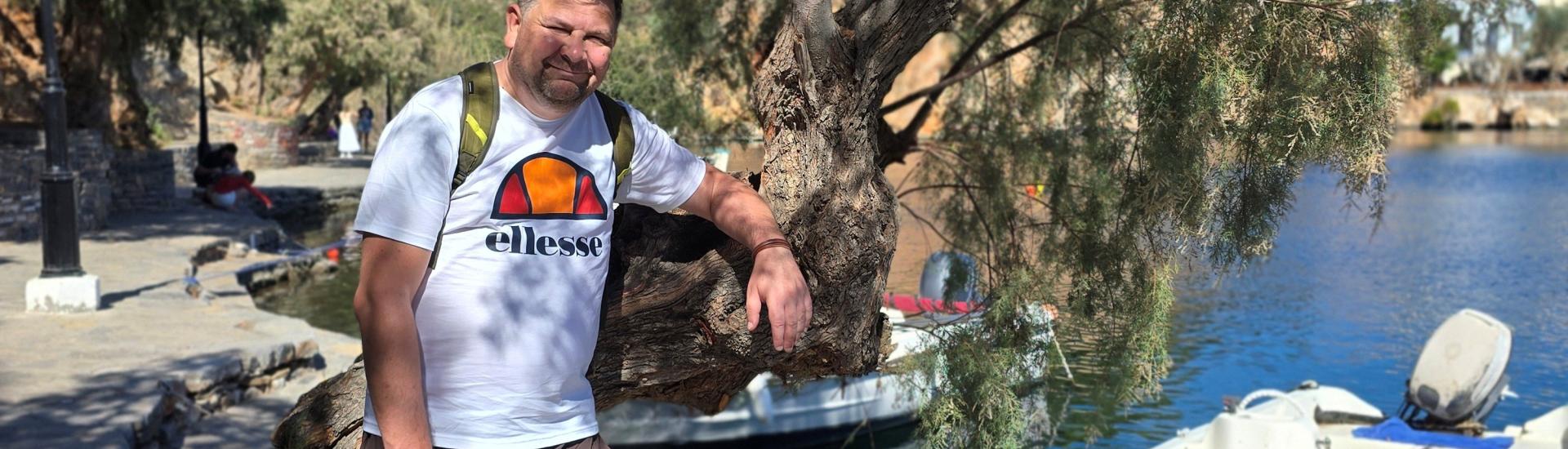 A man stood by a tree in Greece with a boat in the harbour