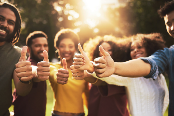 A group of people stand outdoors in a sunlit, natural setting, all giving a thumbs-up gesture toward the camera.