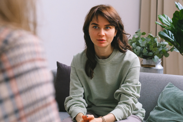 Women sat on a sofa having a conversation with someone. This other person's is facing away from the camera and only their shoulder and back of their head can be seen