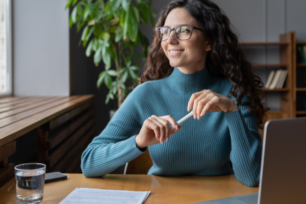 A woman looking out the window whilst sat at a desk and holding a pen